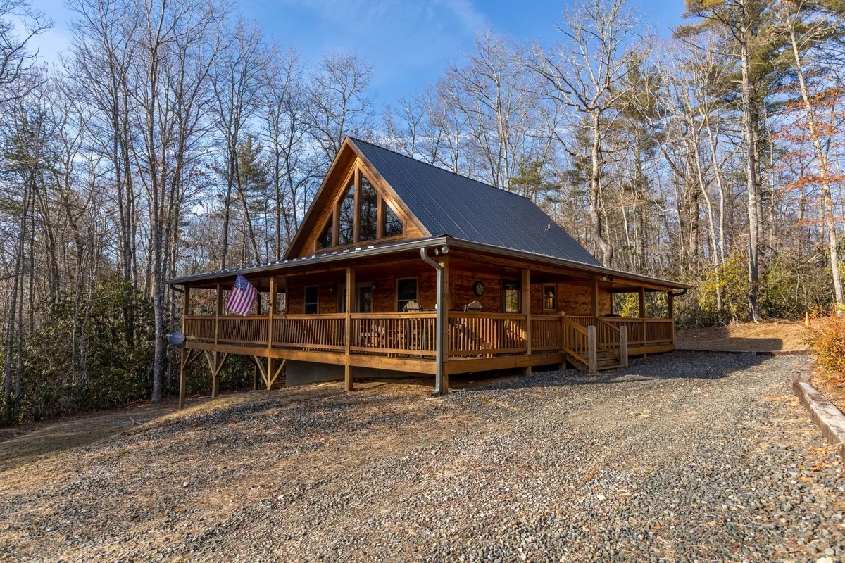 The hero image showcases a charming log cabin surrounded by lush greenery, set against a backdrop of rolling mountains. The spacious front porch, furnished for relaxation, is adorned with an American flag, evoking a sense of warmth and hospitality. The scene suggests tranquil outdoor moments—whether sipping morning coffee, watching sunsets, or gathering with loved ones—all while immersed in the natural beauty of the Blue Ridge Mountains.