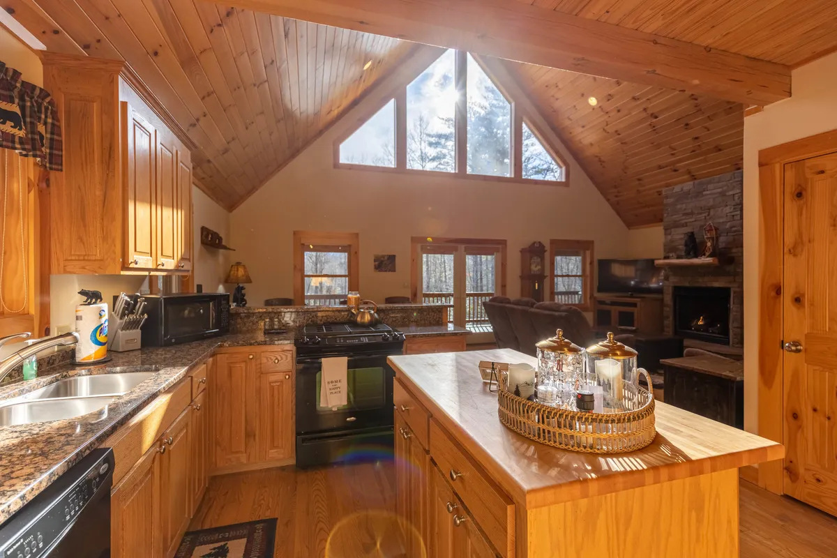 A welcoming kitchen scene with granite countertops, ample wooden cabinetry, and large windows inviting natural light. The central island serves as a hub for socializing, cooking, and shared experiences, while the fireplace in the background hints at continued warmth and togetherness.