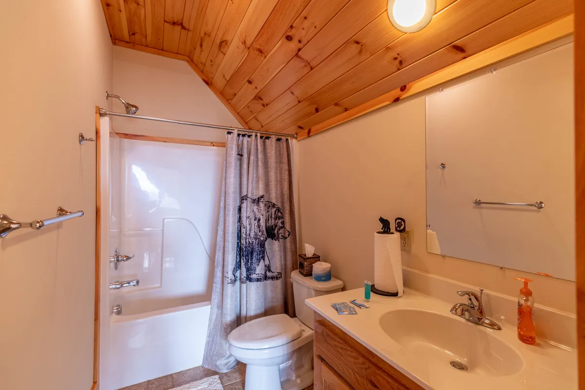 A rustic, warmly-lit bathroom with a wooden ceiling, vanity sink, and a distinctive bear-patterned shower curtain. The compact, comfortable design is perfect for refreshing showers and practical daily routines while staying at the lodge.
