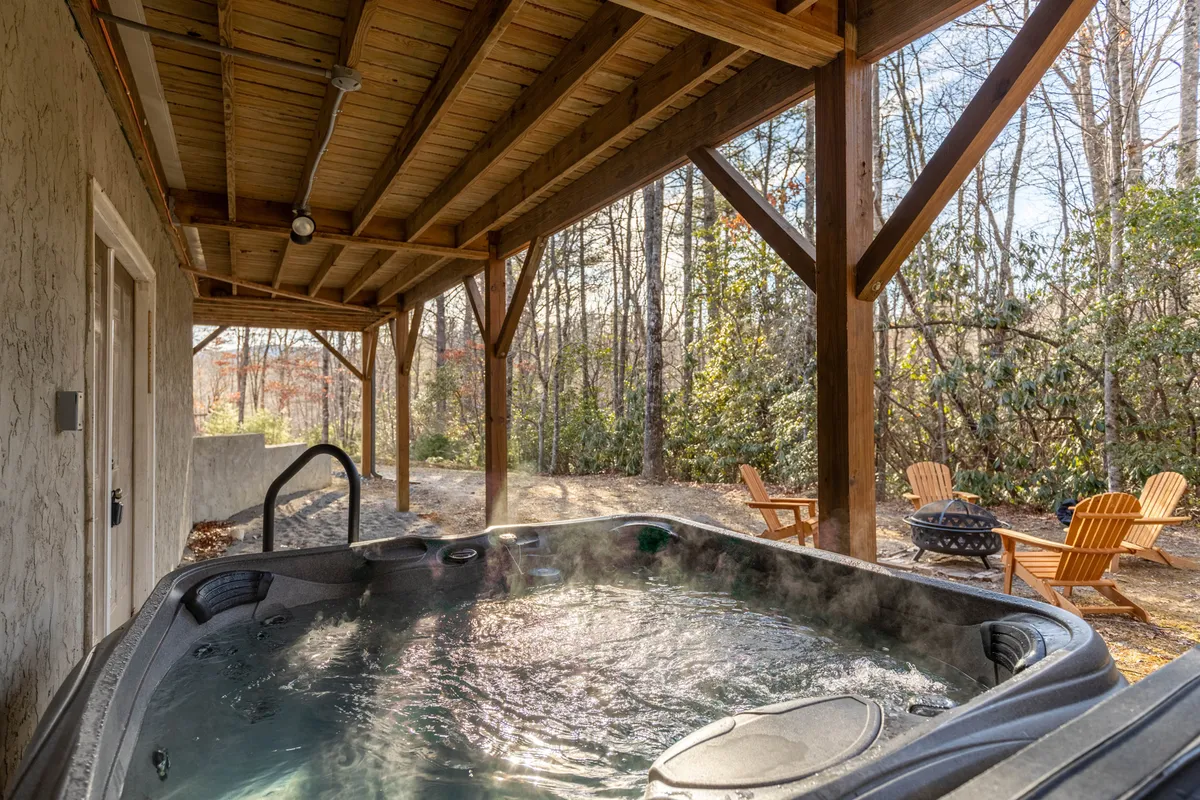 The patio area offers a tranquil setting with a hot tub, wooden seating arrangements, and a fire pit nestled among mature trees. The space feels both private and inviting, ideal for social gatherings, stargazing, or enjoying a peaceful morning coffee surrounded by nature.