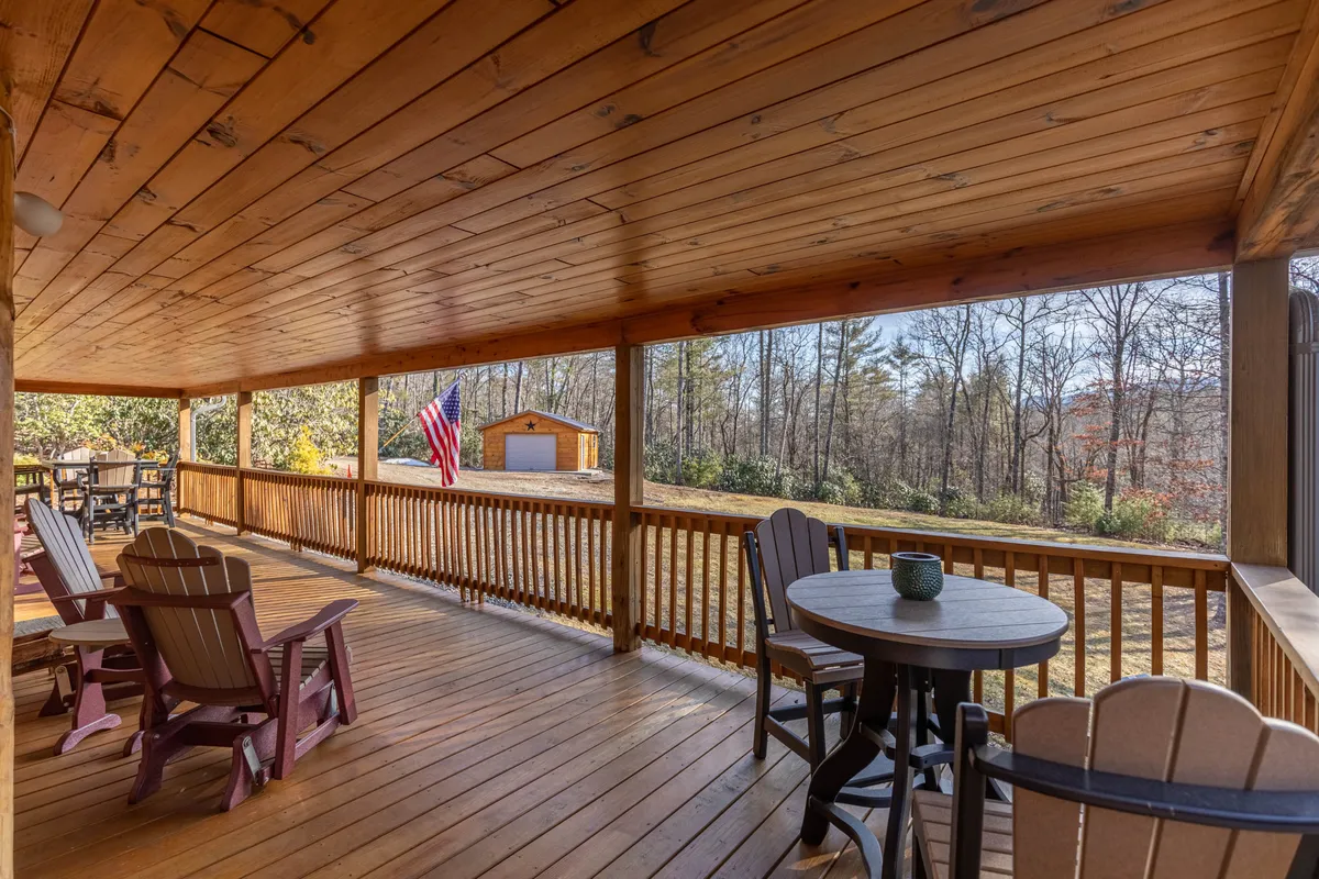 A broad wooden deck features Adirondack chairs and a table adorned with a vase, with an American flag fluttering and a shed in the distance. The grassy yard stretches beyond the deck, offering ample space for kids and pets to play, and the setting is surrounded by lush trees.