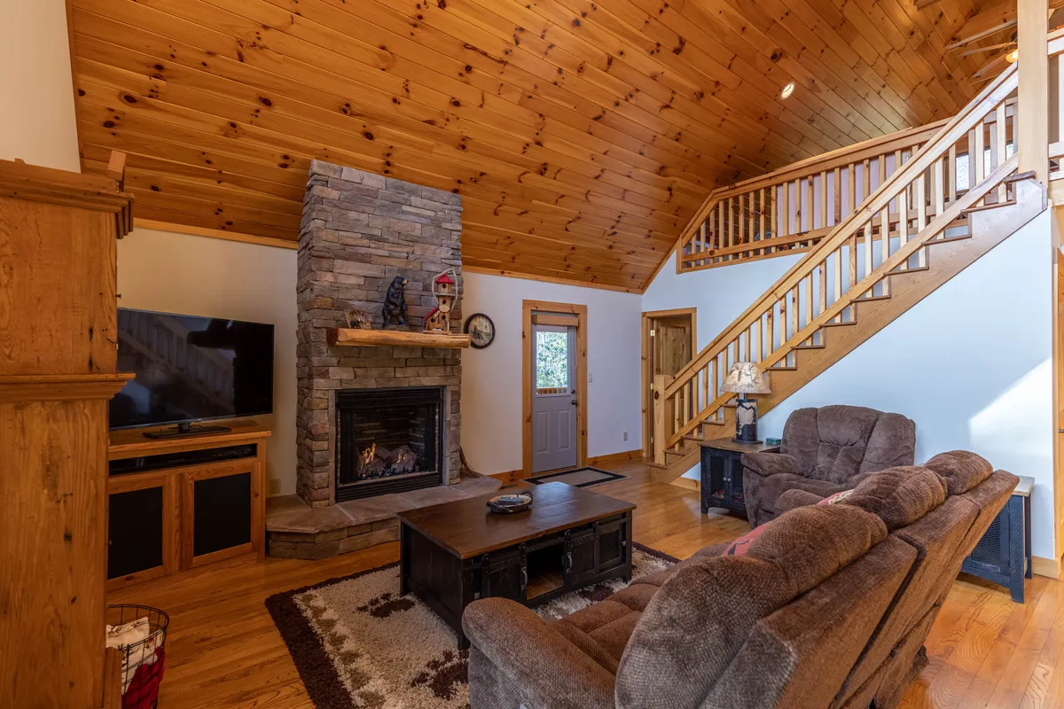This image presents a cozy lodge interior focused on a stone fireplace, wooden staircase, and inviting seating area. The arrangement suggests warmth and togetherness, with elements like a coffee table that encourage group activities such as board games or conversations.