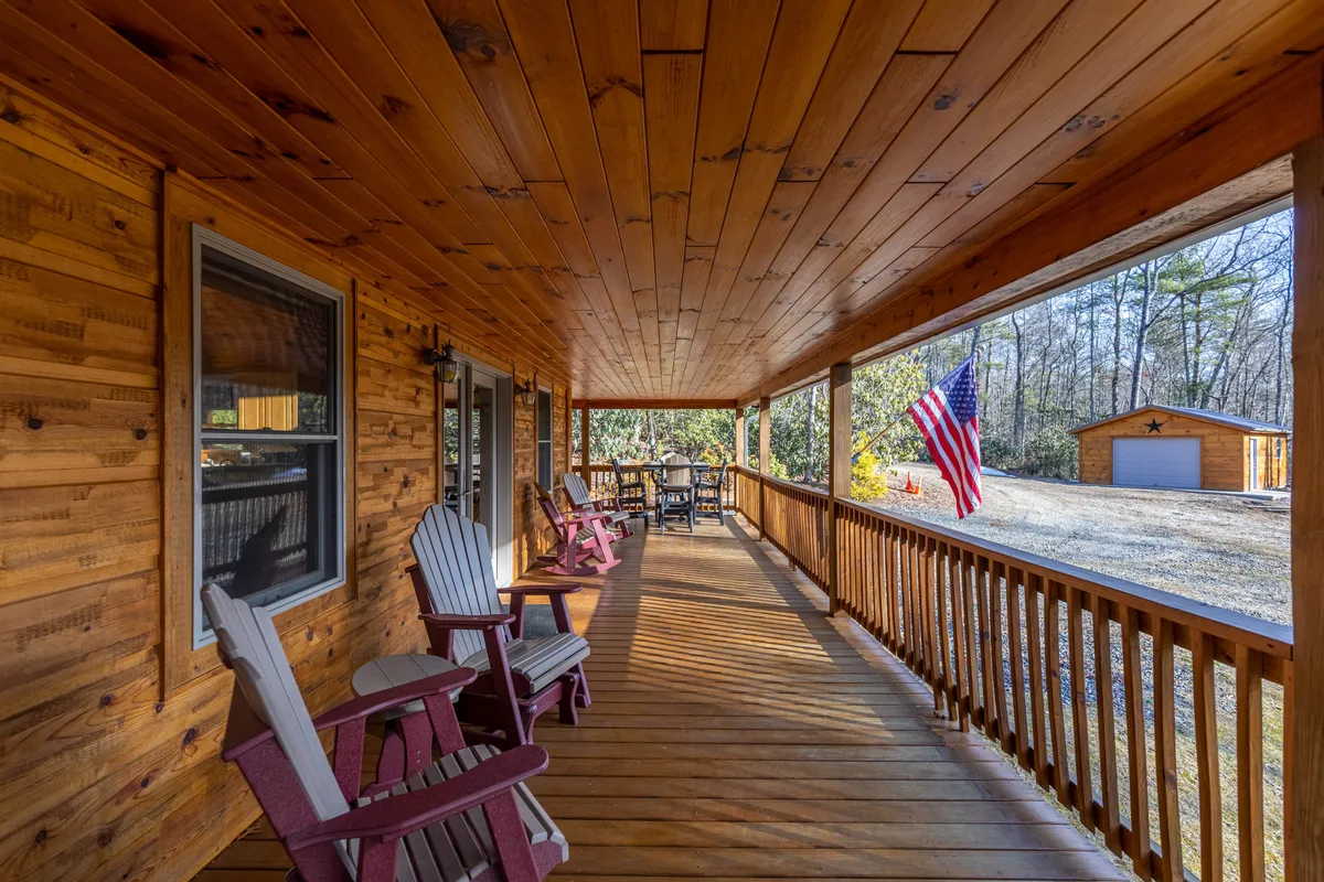 A charming wooden porch surrounded by trees, complete with inviting rocking chairs and an American flag. The setting is enveloped by lush greenery, suggesting privacy and a deep connection to nature. The porch offers a tranquil vantage point for sunsets and relaxed mornings.
