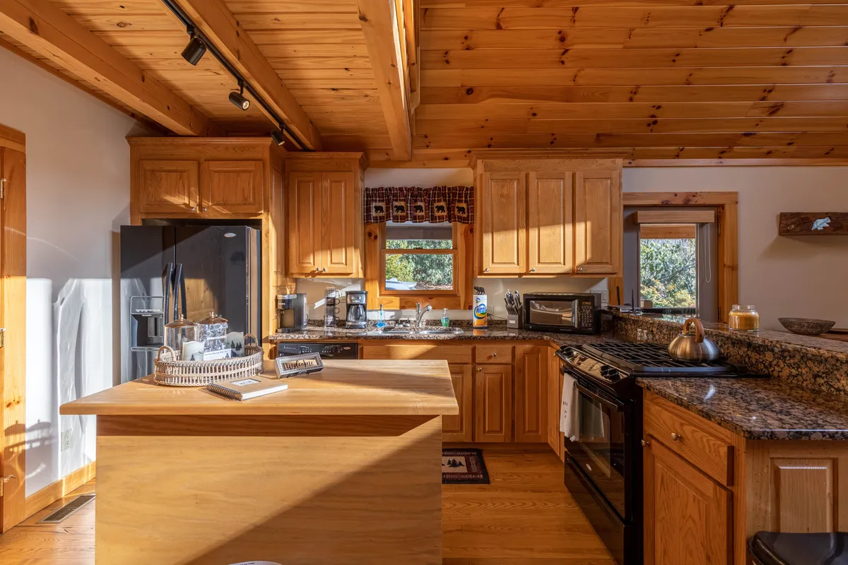 A bright kitchen scene centered around a spacious island with a granite countertop, surrounded by wooden cabinets and bathed in natural light from nearby windows. The island is arranged for social interaction, ideal for group cooking or sharing meals, and the overall ambiance is both cozy and functional.