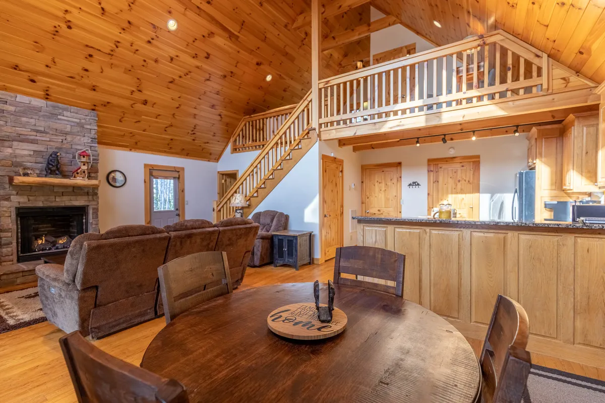A warm, open-concept interior featuring a stone fireplace, wooden dining table, open staircase, and a kitchen area. The arrangement encourages both cooking and relaxing by the fire, with ample space for family gatherings and entertaining guests. The environment exudes a blend of rustic charm and modern convenience.