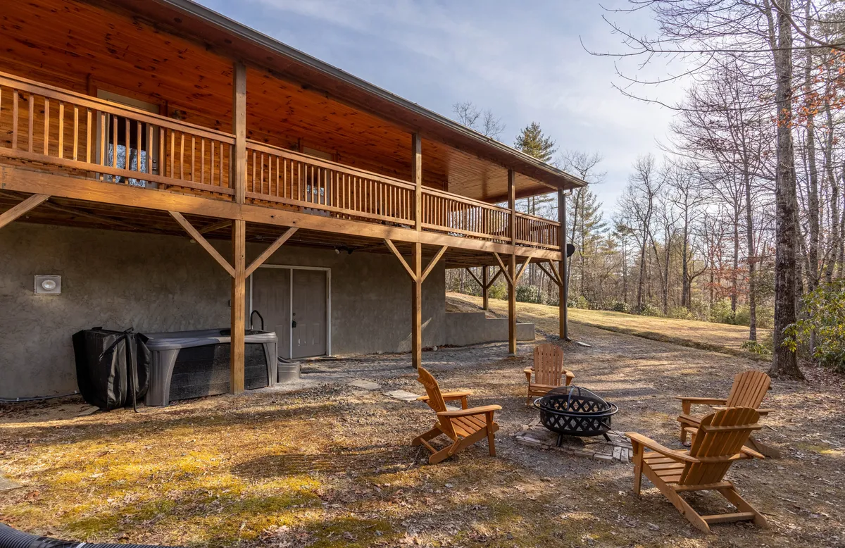 A spacious wooden deck features a cozy fire pit surrounded by Adirondack chairs and a bubbling hot tub in the corner. The area is framed by natural wood railings and lush greenery, creating a seamless blend with the mountain surroundings. Soft evening light hints at sunset, setting a warm, inviting tone perfect for gatherings or quiet relaxation.
