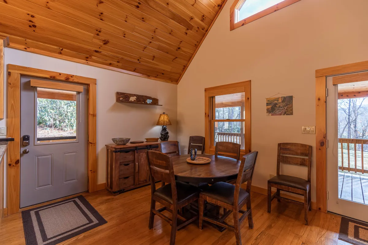 A warm, inviting dining area with a large wooden table and chairs, set beside a substantial window that offers glimpses of the outdoors. Natural light fills the space, and the view through the window hints at the surrounding greenery, reinforcing the lodge’s connection to nature.