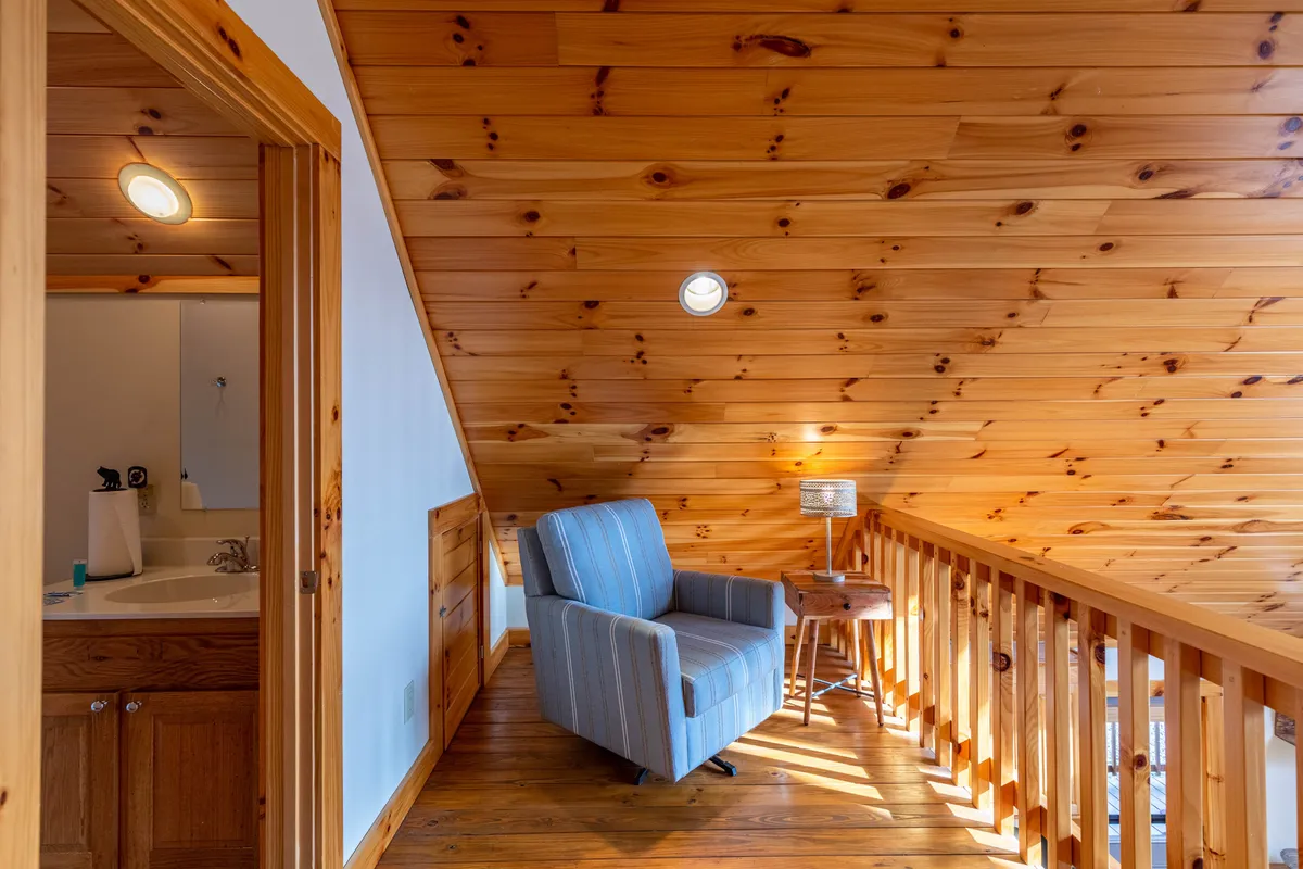 A snug interior featuring an inviting armchair, a wooden railing, a table lamp, and a door leading to a bathroom. The scene suggests a space for quiet relaxation, reading, or conversation, just off the entry, with easy access to a half bath.