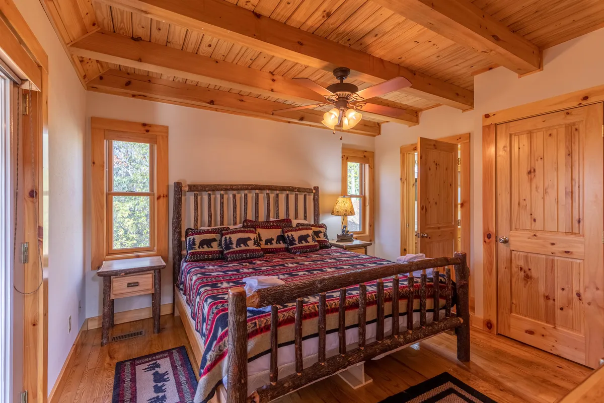 A cozy loft-style bedroom featuring a rustic wooden queen bed, decorative bedding, a nightstand with a lamp, and windows that fill the room with natural light. The space feels both romantic and warm, ideal for couples or guests seeking relaxation.