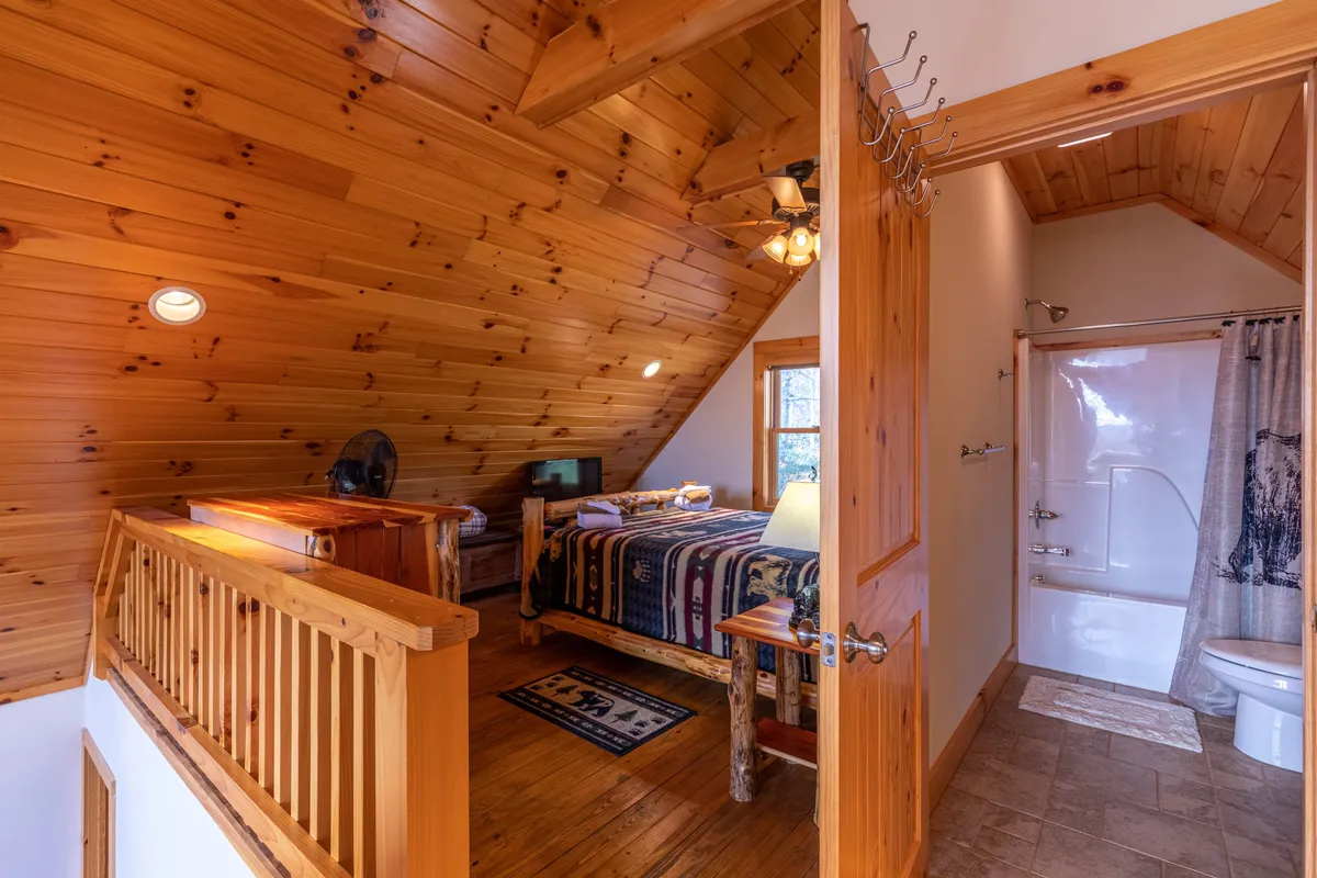 This image captures a rustic bedroom area with log furniture, patterned bedding, and a visible entrance to a bathroom. Natural light fills the space and warm wood tones create a welcoming atmosphere, while the sightline to the bathroom hints at the convenience of attached amenities for comfort during longer stays.