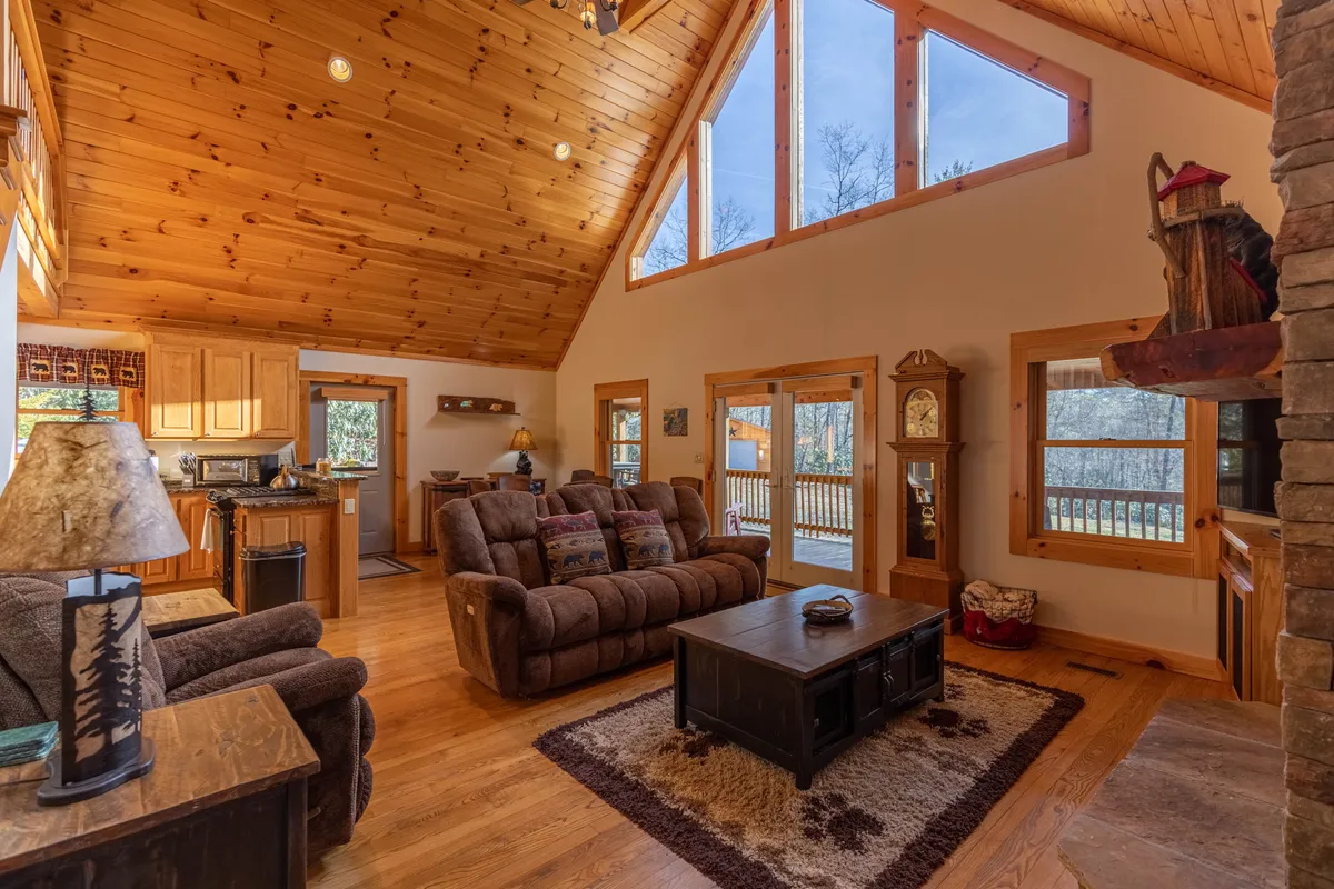 A spacious, open-plan living room and kitchen area with comfortable seating set near a fireplace. The scene includes large windows flooding the space with natural light, a wooden ceiling, and a view into the kitchen area. The layout is designed for both relaxation and social gatherings, with inviting furniture and a blend of modern and rustic touches.