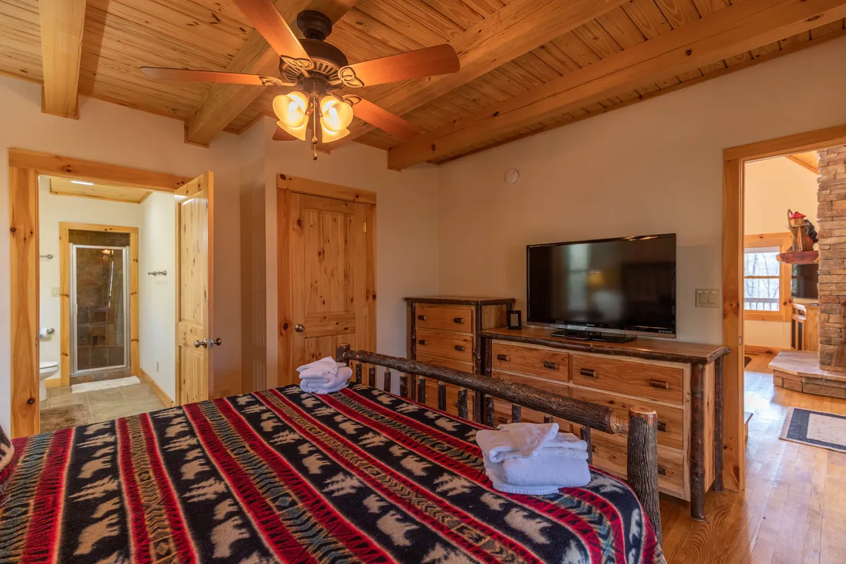 This interior image focuses on a king bed adorned with a patterned blanket, complemented by wooden furniture, a ceiling fan, and a television. Natural light brightens the space, emphasizing comfort and relaxation.